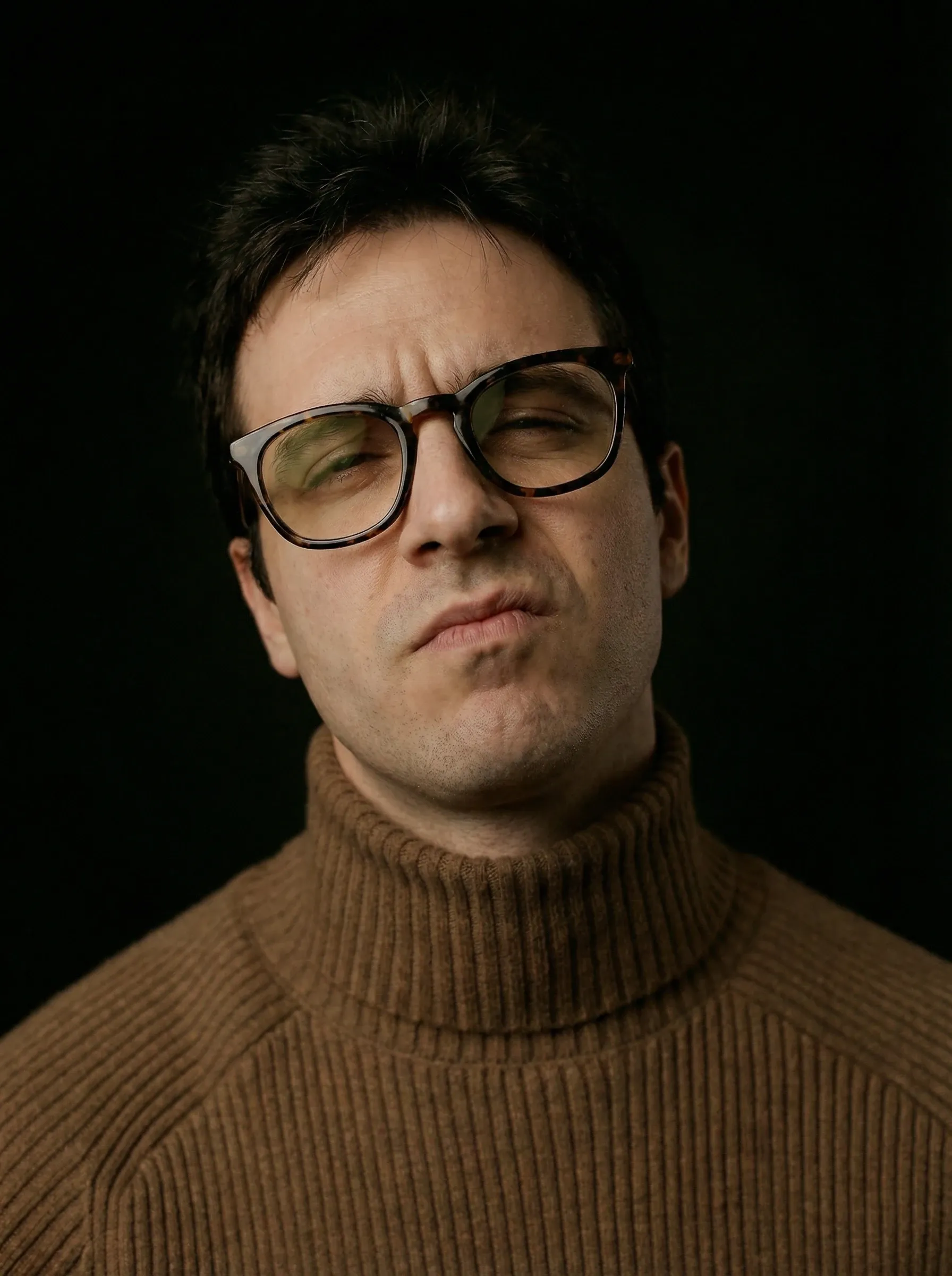 AI actor headshot — man in a brown ribbed turtleneck and glasses against a dark studio background